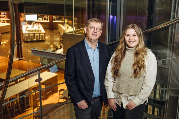 Kaksi henkil&ouml;&auml; seisoo sis&auml;tiloissa lasikaiteen &auml;&auml;rell&auml; modernissa toimisto- tai tapahtumaymp&auml;rist&ouml;ss&auml;. Taustalla n&auml;kyy avoin aulatila ja valaistuja rakenteita.